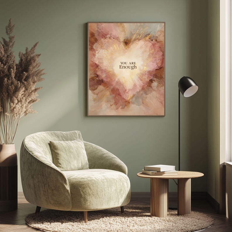 May include: A framed artwork with a heart-shaped design in shades of pink, gold, and beige, with the words "YOU ARE Enough" in the center. The artwork hangs on a green wall above a green velvet armchair. A small wooden side table and a floor lamp are also in the room.