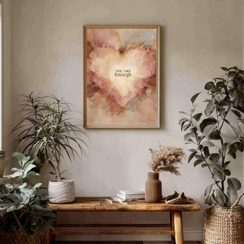 May include: A framed art print featuring a heart-shaped design in shades of pink, gold, and beige. The words "YOU ARE Enough" are centered within the heart. The artwork is displayed above a wooden bench with plants.