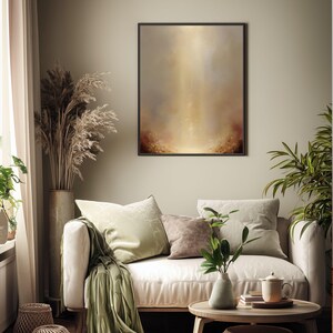 May include: A framed abstract art print with a warm color palette hangs above a cream-colored sofa. The artwork features a blend of gold, brown, and beige tones, creating a soft, ethereal effect. Decorative pillows and a green throw blanket add texture.