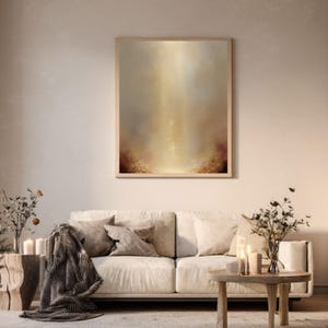 May include: Abstract art print in a wooden frame. The artwork features a warm color palette of beige, gold, and brown, with a soft, blurred effect. The painting is displayed above a cream-colored sofa with throw pillows and a blanket.
