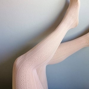 Peut inclure: Une paire de collants en dentelle blanche avec un motif délicat. Les collants sont portés sur les jambes d'une personne, une jambe levée et l'autre pliée au niveau du genou.