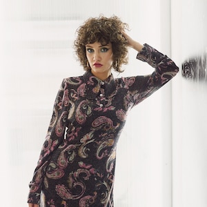 May include: A woman with curly brown hair is wearing a black dress with a paisley print. The dress has long sleeves and a collared neckline. She is wearing white tights with a small white bow pattern.