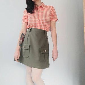 May include: A vintage-style outfit featuring a short-sleeved orange and white gingham shirt and an olive green mini skirt with two front pockets. The skirt has gold-colored button details. The outfit is paired with white tights.