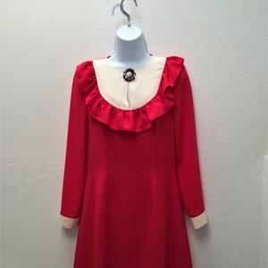 May include: A red dress with a white collar and ruffled trim. The dress has long sleeves and a jewel-toned brooch at the neckline.