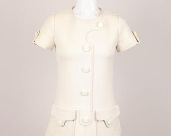 May include: Ivory-colored dress with short sleeves, a round neckline, and a-line skirt. The dress features a series of large, round buttons down the front and two pockets with button details. The dress is displayed on a mannequin.