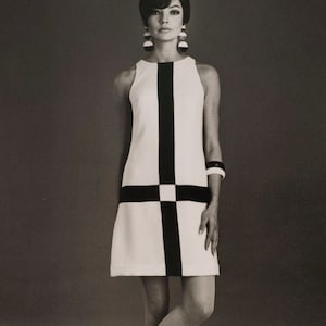 May include: A woman wearing a white and black geometric patterned sleeveless dress with a black belt. She is wearing large round earrings and a black bracelet.