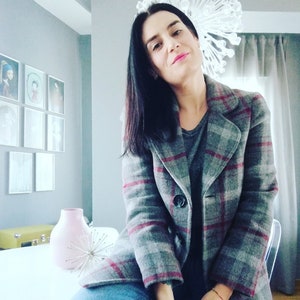 Puede incluir: Una mujer con una chaqueta de cuadros grises y rojos con cierre de bot&oacute;n. Est&aacute; sentada en una silla blanca con las piernas cruzadas. Tiene el pelo largo y negro y lleva pantalones vaqueros azules.