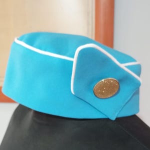 May include: A turquoise hat with white edging and a gold-coloured button. The hat is rectangular with a curved top and a small side flap. It is likely a uniform accessory, possibly for a flight attendant.