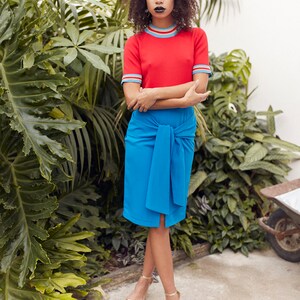 May include: A woman wearing a red short-sleeved shirt with blue and white stripes around the neck and sleeves, a blue skirt tied at the waist, and gold sandals. She is standing in front of a wall of green plants.