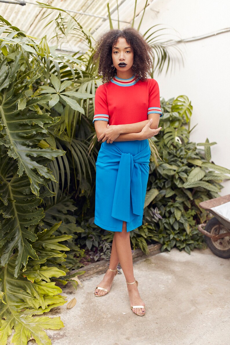 May include: A woman wearing a red short-sleeved shirt with blue and white stripes around the neck and sleeves, a blue skirt tied at the waist, and gold sandals. She is standing in front of a wall of green plants.