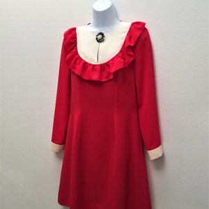 May include: A red dress with a white ruffle collar and long sleeves. The dress has a white trim around the collar and cuffs. A small jeweled brooch is attached to the collar.