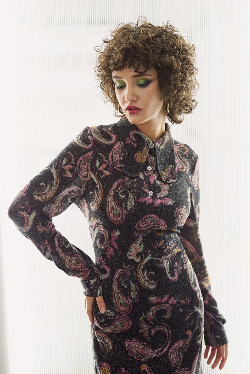 May include: A woman with curly brown hair wears a black and pink paisley patterned dress with a collared neckline and long sleeves.