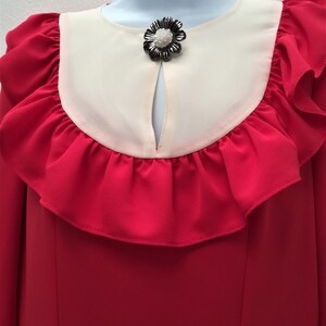 May include: A red dress with a white ruffle collar and a black flower brooch. The dress has long sleeves and a fitted bodice.
