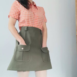 May include: A vintage-style outfit featuring a short-sleeved orange and white gingham shirt paired with an olive green skirt. The skirt has two front pockets with decorative buttons. The outfit is a retro fashion statement.