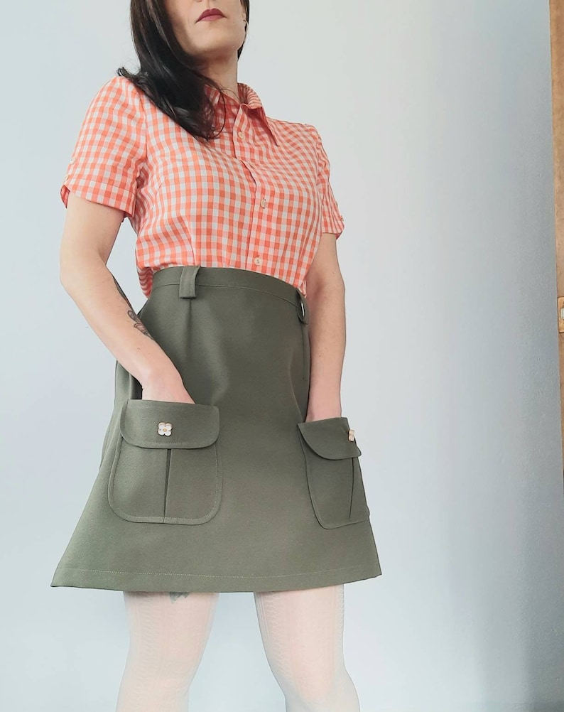 May include: A vintage-style outfit featuring a short-sleeved orange and white gingham shirt paired with an olive green skirt. The skirt has two front pockets with decorative buttons. The outfit is a retro fashion statement.