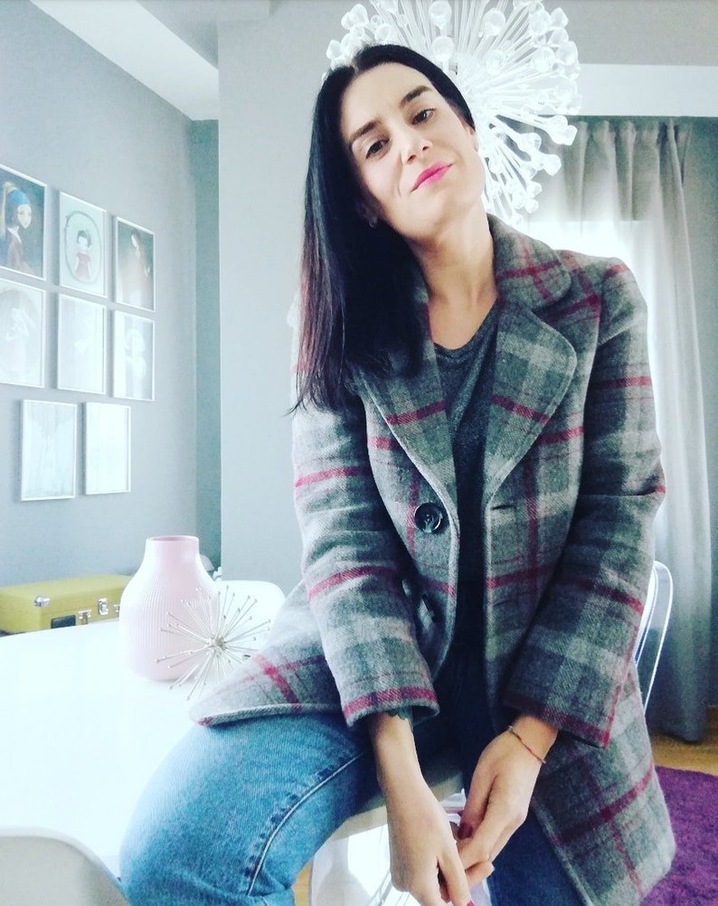 Puede incluir: Una mujer con una chaqueta de cuadros grises y rojos con cierre de bot&oacute;n. Est&aacute; sentada en una silla blanca con las piernas cruzadas. Tiene el pelo largo y negro y lleva pantalones vaqueros azules.