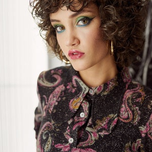 May include: A woman with curly brown hair wearing a black and pink paisley patterned shirt with gold hoop earrings. She has green and blue eyeshadow and pink lipstick.