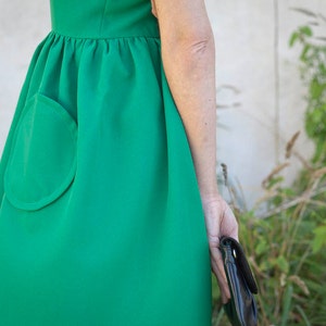 K&ouml;nnte beinhalten: Ein leuchtend gr&uuml;nes Kleid mit einer gro&szlig;en, abgerundeten Tasche auf der linken Seite. Das Kleid hat eine geraffte Taille und einen ausgestellten Rock.