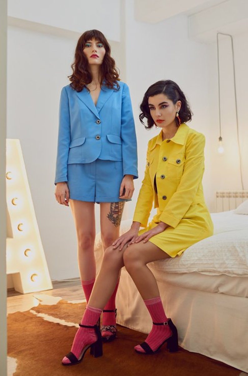May include: Two women wearing matching pink socks and black heels. One woman is wearing a light blue blazer and shorts, the other is wearing a yellow blazer and skirt.