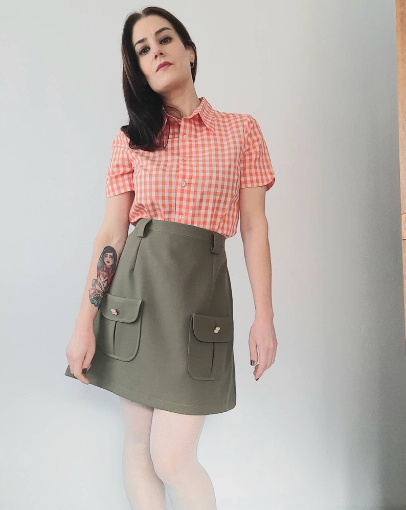 May include: A vintage-style outfit featuring a short-sleeved orange and white gingham shirt and an olive green mini skirt with two front pockets. The skirt has gold-colored button details. The outfit is paired with white tights.