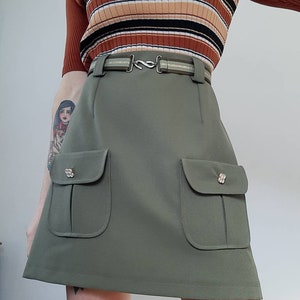 May include: Olive green mini skirt with two front pockets and decorative buttons. The skirt has a matching belt with a silver-tone buckle. The model is wearing a brown, striped, ribbed top.