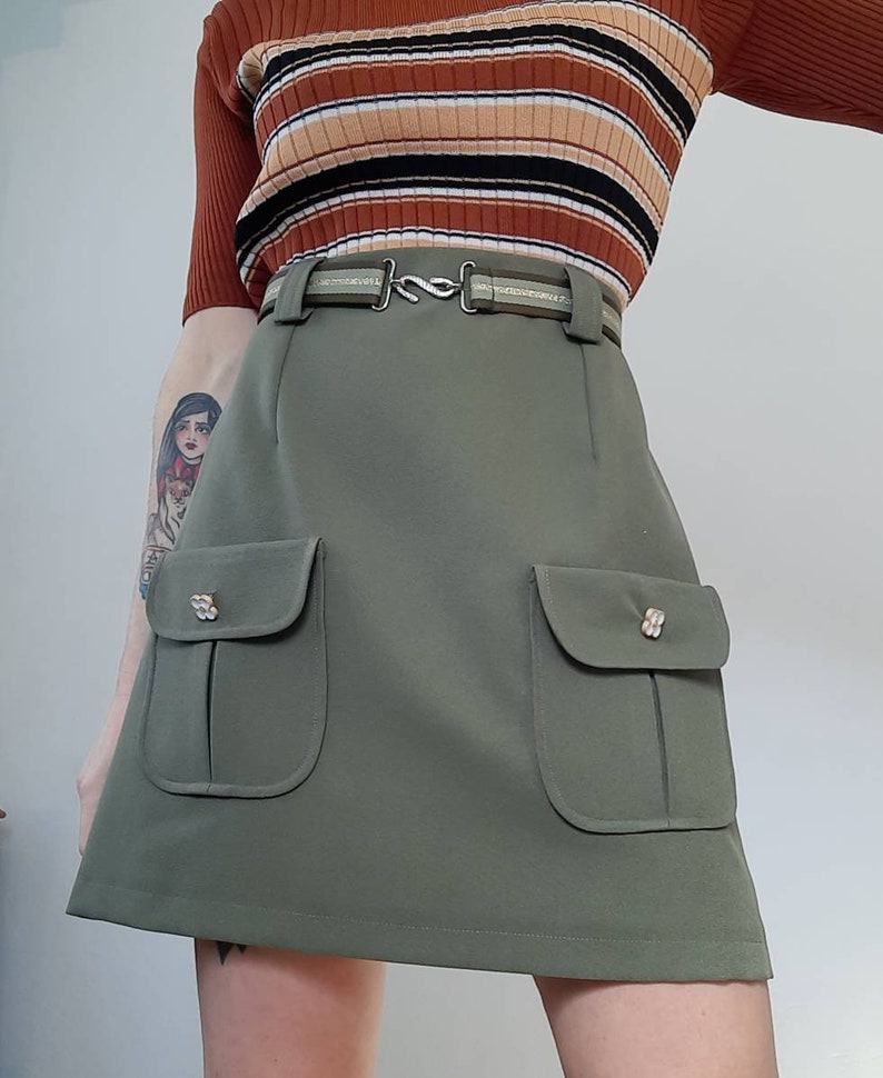 May include: Olive green mini skirt with two front pockets and decorative buttons. The skirt has a matching belt with a silver-tone buckle. The model is wearing a brown, striped, ribbed top.
