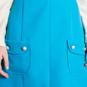 May include: A turquoise blue skirt with two large pockets and silver buttons. The skirt is a-line and has a simple design.
