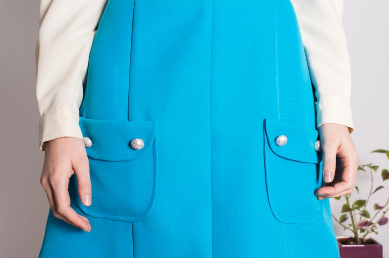 May include: A turquoise blue skirt with two large pockets and silver buttons. The skirt is a-line and has a simple design.