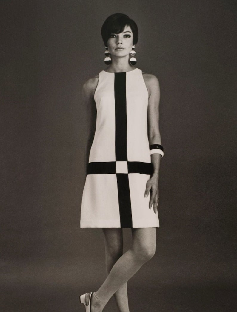 May include: A woman wearing a white and black geometric patterned sleeveless dress with a black belt. She is wearing large round earrings and a black bracelet.