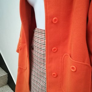 May include: A bright orange coat with a double breasted closure and a large patch pocket on the right side. The coat is worn over a plaid skirt.
