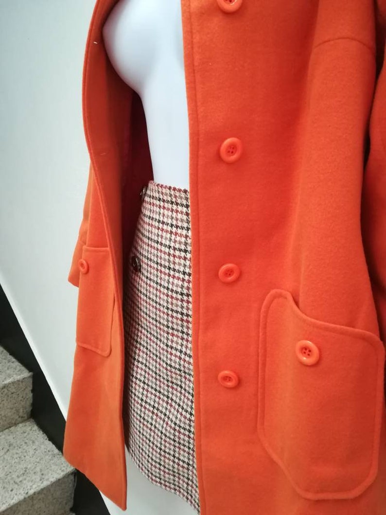 May include: A bright orange coat with a double breasted closure and a large patch pocket on the right side. The coat is worn over a plaid skirt.