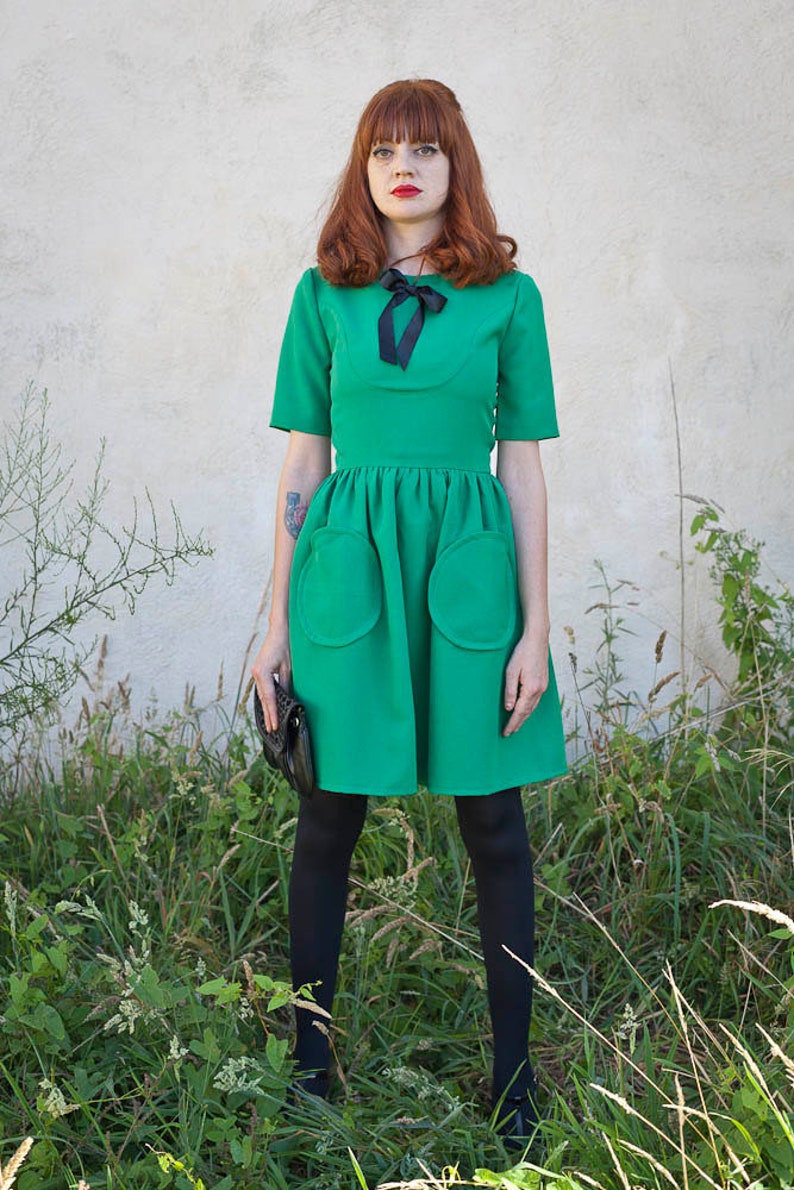 K&ouml;nnte beinhalten: Eine Frau in einem gr&uuml;nen Kleid mit einer schwarzen Schleife und schwarzen Strumpfhosen. Sie steht auf einer gr&uuml;nen Wiese.