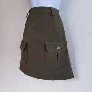 May include: Olive green mini skirt with two front pockets and decorative gold-colored buttons. The skirt has a high waist and a belt loop. The fabric appears to be a textured weave.