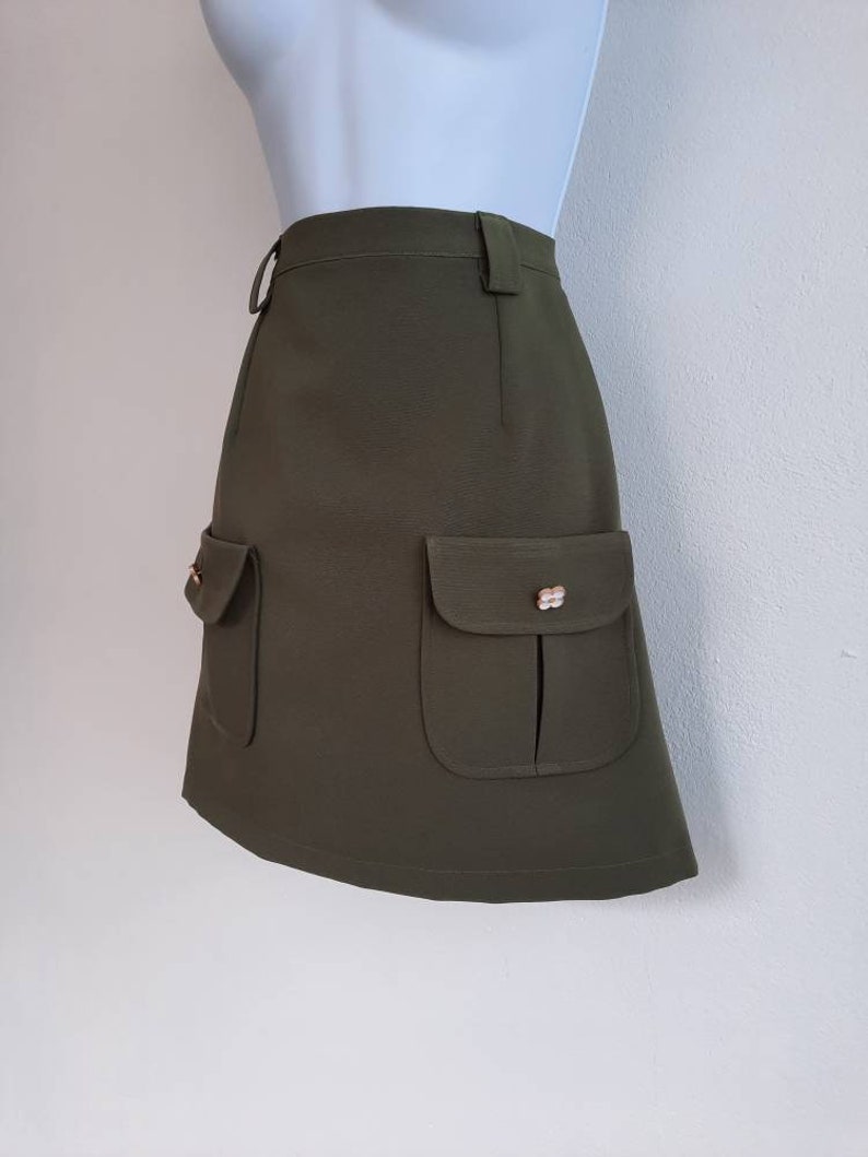 May include: Olive green mini skirt with two front pockets and decorative gold-colored buttons. The skirt has a high waist and a belt loop. The fabric appears to be a textured weave.