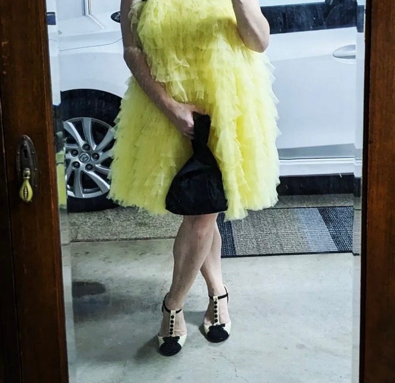 以下が含まれることがあります： 黄色のチュールスカートのドレスと黒いハイヒールを履いた女性。黒いクラッチバッグを持っています。