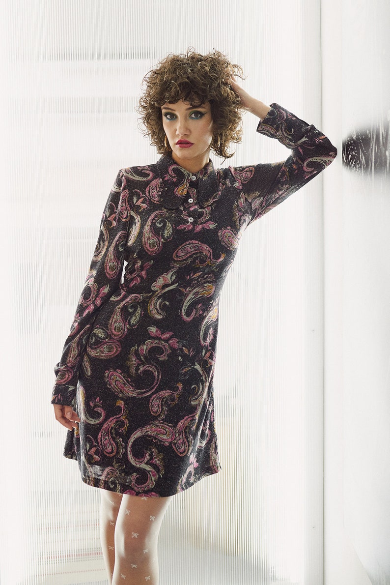 May include: A woman with curly brown hair is wearing a black dress with a paisley print. The dress has long sleeves and a collared neckline. She is wearing white tights with a small white bow pattern.