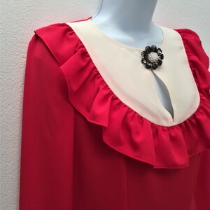 May include: A red blouse with a white collar and a black flower brooch. The blouse has a ruffled neckline and long sleeves.