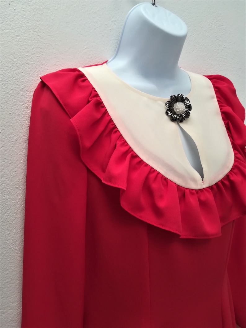 May include: A red blouse with a white collar and a black flower brooch. The blouse has a ruffled neckline and long sleeves.