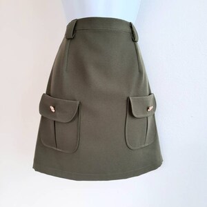 May include: Olive green skirt with two front pockets and decorative gold-colored buttons. The skirt has a tailored fit and a classic design, suitable for various occasions. The skirt is displayed on a mannequin.