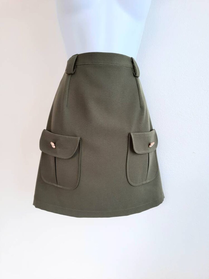 May include: Olive green skirt with two front pockets and decorative gold-colored buttons. The skirt has a tailored fit and a classic design, suitable for various occasions. The skirt is displayed on a mannequin.