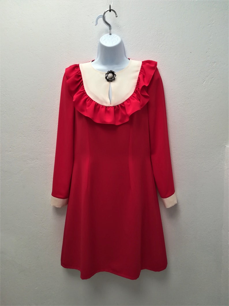 May include: A red dress with a white collar and ruffled trim. The dress has long sleeves and a jewel-toned brooch at the neckline.