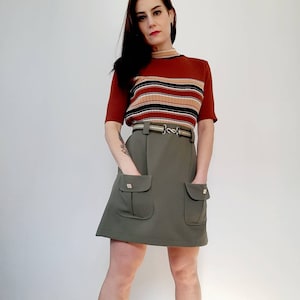 May include: A woman wearing a vintage outfit. The outfit includes a rust-colored, short-sleeved sweater with horizontal stripes and an olive green skirt with two front pockets. The skirt is paired with a belt.