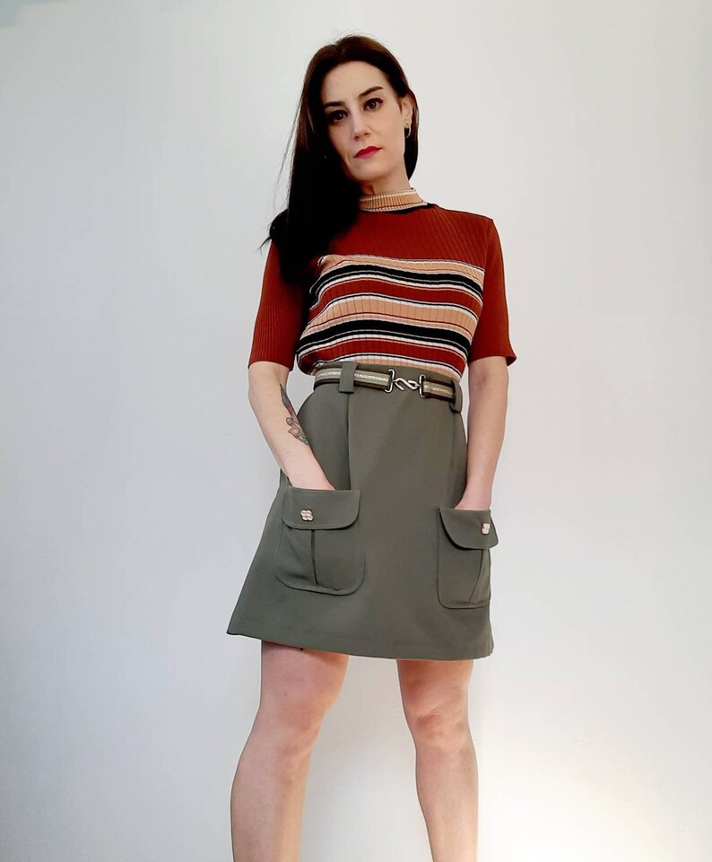 May include: A woman wearing a vintage outfit. The outfit includes a rust-colored, short-sleeved sweater with horizontal stripes and an olive green skirt with two front pockets. The skirt is paired with a belt.