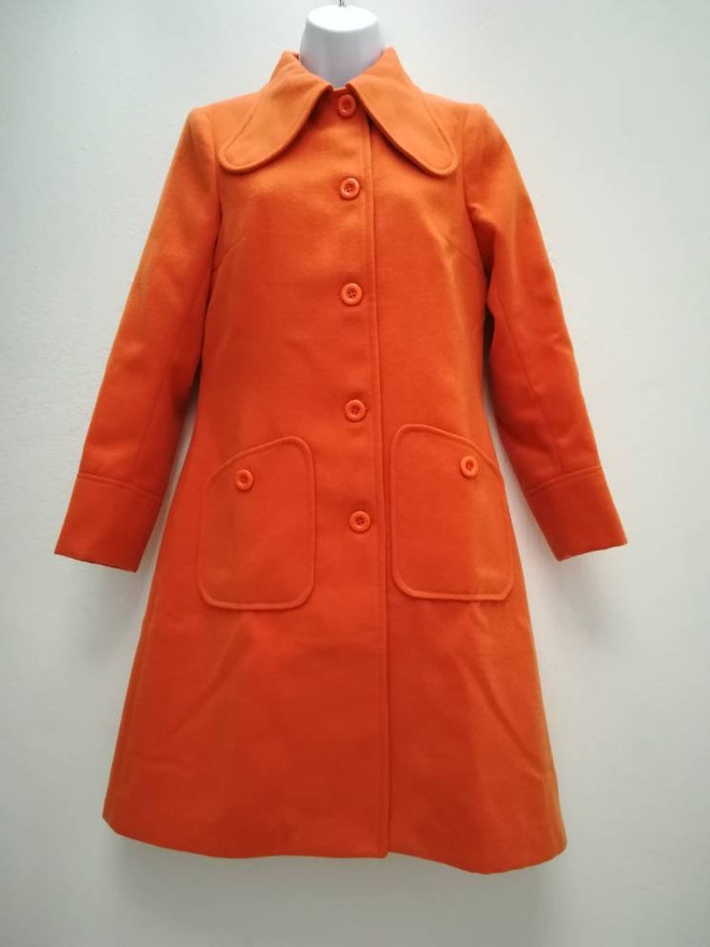 May include: A long-sleeved orange coat with a large collar and a button closure. The coat has two large pockets on the front.