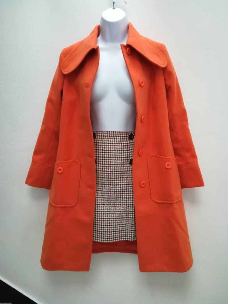 May include: A bright orange coat with a large collar and two pockets. The coat is on a white mannequin and has a button closure.