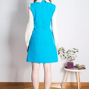 May include: A woman wearing a blue dress with a white collar and white sleeves. The dress is a-line and has a zipper in the back. She is wearing white tights with black polka dots and black heels.