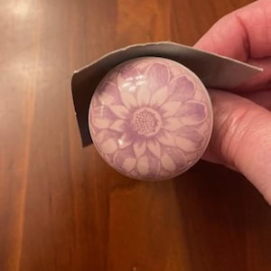 May include: A round, pale purple cabinet knob featuring a detailed floral pattern in a darker shade of purple. The knob is partially covered by a grey cardboard backing. The knob is likely made of ceramic or porcelain.