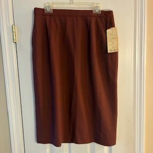 Könnte beinhalten: Ein bordeauxfarbener Bleistiftrock hängt an einer weißen Tür. Der Rock hat einen Knopfverschluss und ein angebrachtes Etikett. Der Rock ist aus einem glatten Stoff und hat eine einfarbige Farbe. Der Rock hängt an einem durchsichtigen Kunststoffbügel.