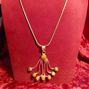 May include: A silver-toned pendant necklace with a unique design. The pendant features a central, round, olive-green stone, with multiple curved silver lines extending downwards, each ending in a smaller, similar-colored stone. The necklace is displayed on a red velvet surface.