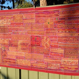 Pode incluir: Um grande tecido retangular com um design de patchwork. O tecido apresenta uma mistura vibrante de tecidos vermelhos, laranja e dourados, com padrões bordados intrincados e uma borda vermelha. O tecido está pendurado em uma cerca verde.